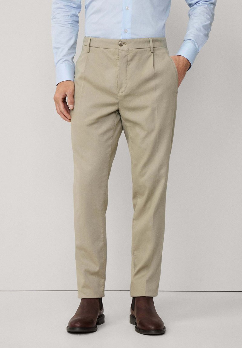 Beige Chinos mit taillierter Passform, aus Baumwolle, mit Vordertaschen und Knopfverschluss. Kombiniert mit braunen Ankle Boots.