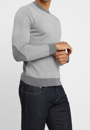 Mies, joka pukeutunut vaaleanharmaaseen teksturoituun neulepuseroon, jossa on tummemmat harmaat resorit ja kyynärpäiden tahrat, yhdistettynä tummansinisiin farkkuihin.