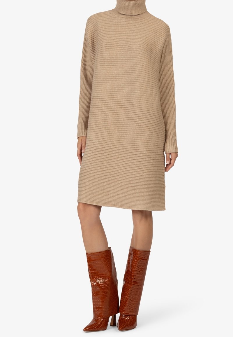 Robe pull en maille côtelée beige avec un col roulé haut, associée à des bottes marron brillantes à motif crocodile montantes jusqu'au genou.