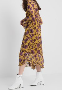 Žluté květinové midi šaty s fialovými vzory, průhledná látka a volánkový lem; nošeny s bílými kotníkovými botami na blokovém podpatku.