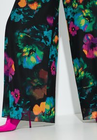 Průsvitné kalhoty s květinovým potiskem na černém pozadí, s živými barvami jako růžová, zelená a oranžová, doplněné fuchsiovými vysokými podpatky.
