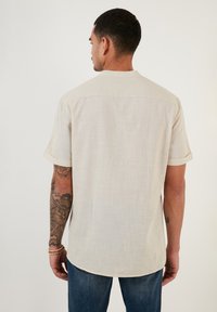 Cămașă scurtă cu mânecă scurtă, de culoare bej deschis, dintr-un material texturat, cu spatele tip yoke și mâneci răsucite. Dispune de o margine curbată și un design simplu.