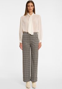 Lichtbeige blouse met een geknoopte strikdetail, gecombineerd met grijs geruite wijde broek, afgewerkt met lichtgekleurde blokhakken.