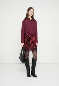 Blusa bordeaux con bottoni abbinata a una gonna rossa con paillettes e rifiniture in pizzo, stivali neri alti fino al ginocchio e una borsa tote nera.