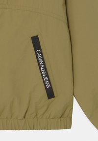 Olivgrüner Windbreaker aus strukturiertem Material, mit elastischem Saum und einer Seitentasche, die ein schwarzes Etikett mit der Aufschrift "CALVIN KLEIN JEANS" trägt.