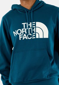 Sudadera pullover en color azul verdoso con un bolsillo tipo canguro en la parte delantera y texto de logo en blanco "THE NORTH FACE" con un diseño arqueado. Textura de tela suave.