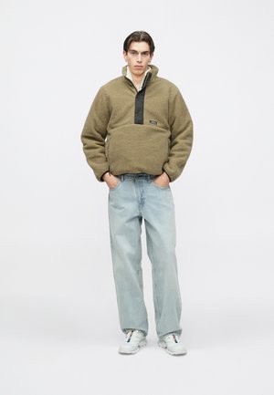 Olivgrüner Fleecepullover mit schwarzem Einsatz und Logopatch; kombiniert mit hellblauen, gerade geschnittenen Jeans und weißen Turnschuhen.