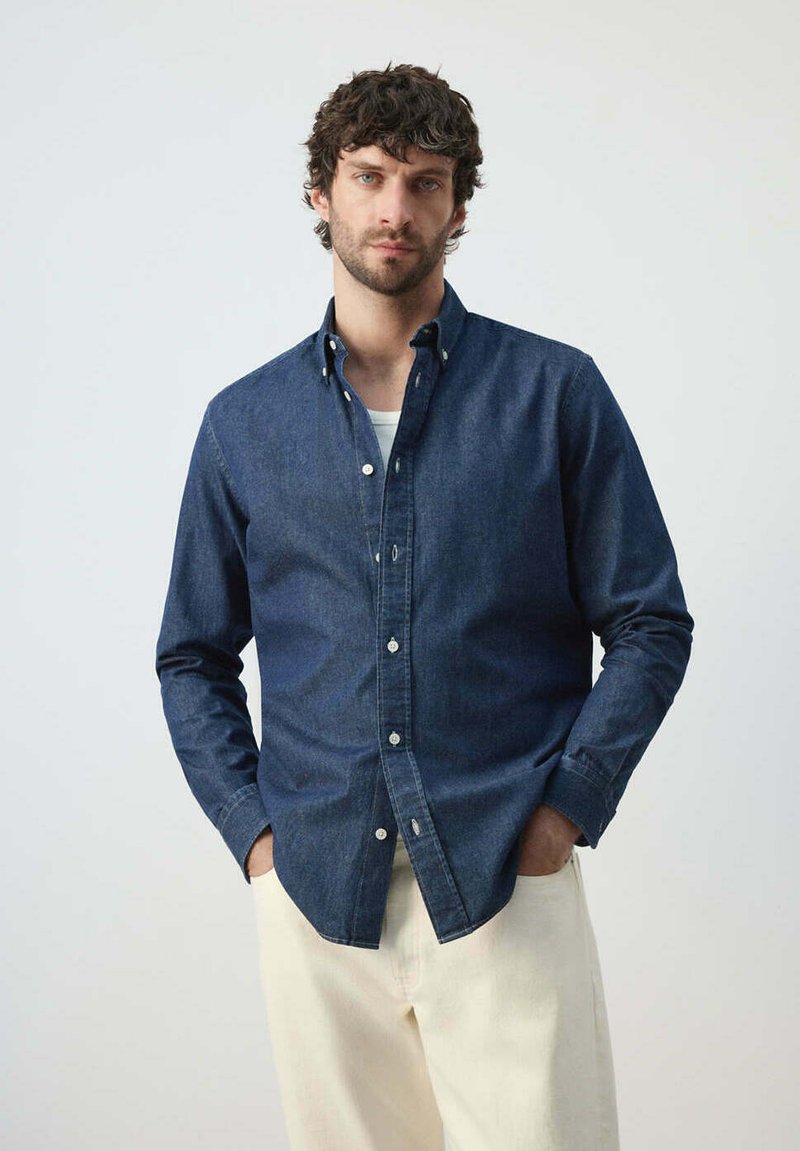 Homme aux cheveux courts et bouclés portant une chemise bleu foncé boutonnée par-dessus un sous-vêtement blanc et un pantalon beige clair, debout les mains dans les poches.