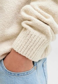 Šviesiai smėlinis megztinis su tekstūrine, pūkuota paviršiumi ir raukšlėtais rankogaliais, derinamas su šviesiai mėlynomis džinsomis, demonstruojantis laisvą siluetą.