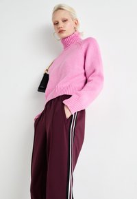 Pink knitted turtleneck sweater with ribbed hem, paired with burgundy trousers featuring white side stripes and side pockets. Accessories include a small black bag.