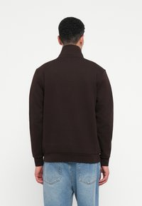 Άνδρας που φοράει μια σκούρα καφέ φούτερ με ψηλό γιακά και ανοιχτό γαλάζιο τζιν, με την πλάτη στραμμένη προς μια απλή λευκή γέφυρα.