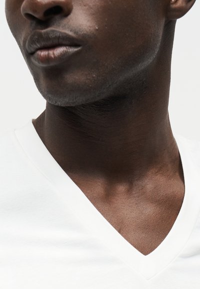 Κοντινό πλάνο του κάτω μέρους του προσώπου, του λαιμού και της κλείδας ενός σκουρόχρωμου άνδρα που φοράει λευκό μπλουζάκι V-neck σε απλό φόντο.