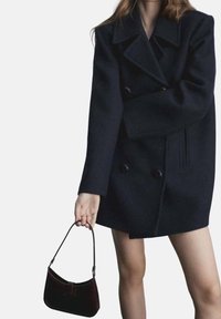 Tummansininen villasta valmistettu kaksirivinen takki, jossa on suuret kaulukset ja sivutaskut, yhdistettynä pieneen, kaarevaan mustaan käsilaukkuun. Sileä pinta, rakenteellinen muotoilu.