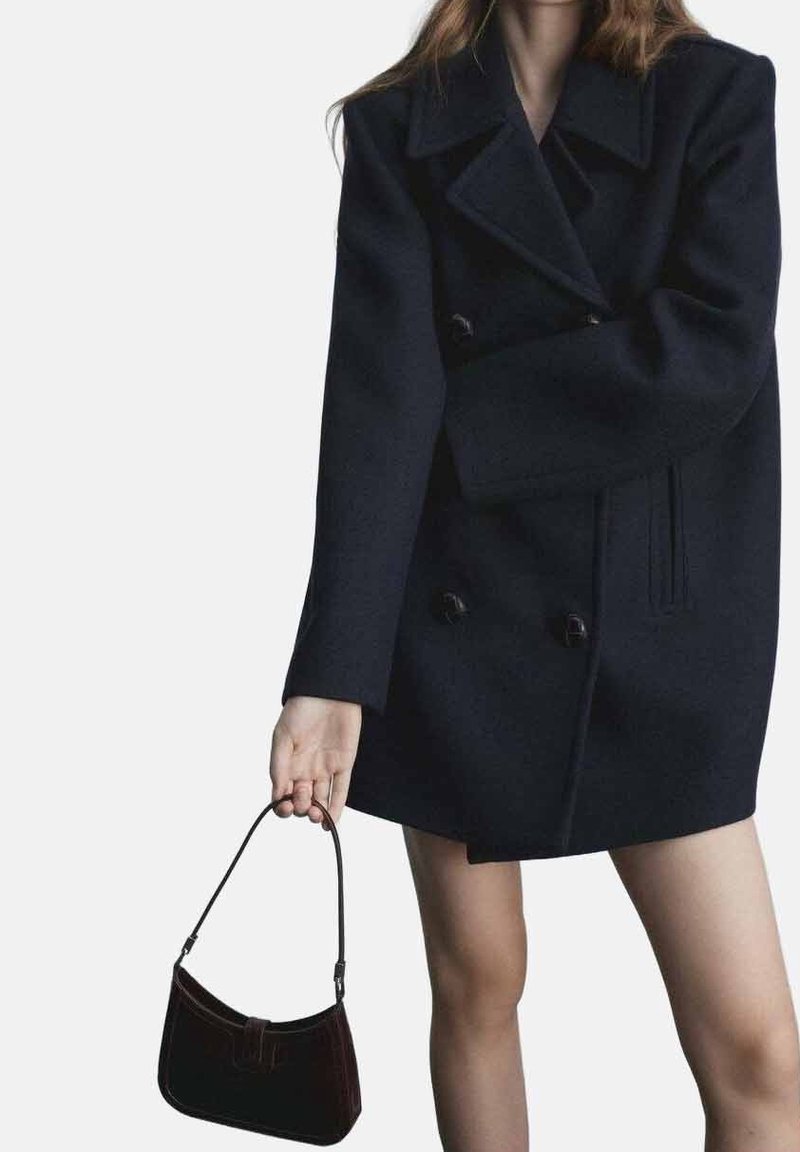 Tummansininen villasta valmistettu kaksirivinen takki, jossa on suuret kaulukset ja sivutaskut, yhdistettynä pieneen, kaarevaan mustaan käsilaukkuun. Sileä pinta, rakenteellinen muotoilu.