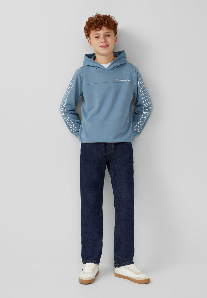 Lichtblauwe hoodie met witte tekst op de voorkant en mouwen, gecombineerd met donkerblauwe jeans en witte sneakers met tan kleurige zolen.