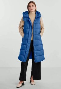 Mėlynas pūkinis liemelis su kapucu, turintis užtrauktuką ir spaudę, su siuvinėta tekstūra ir šoninėmis kišenėmis, derinamas su kreminiu megztiniu ir juodomis kelnėmis.