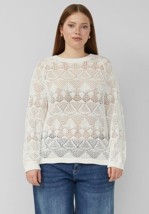 Kobieta z długimi włosami, nosząca złote kolczyki koła, biały sweter w wzory i niebieskie dżinsy, stojąca na tle szarego tła.