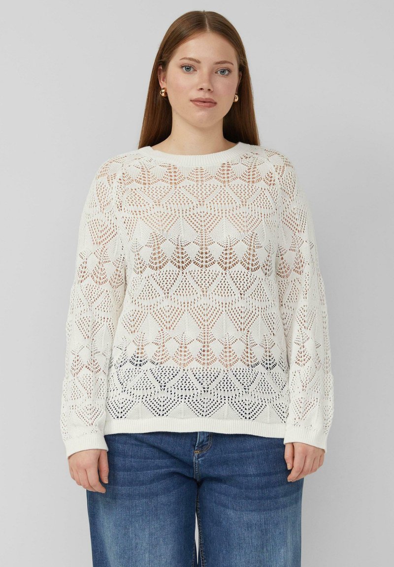 Kobieta z długimi włosami, nosząca złote kolczyki koła, biały sweter w wzory i niebieskie dżinsy, stojąca na tle szarego tła.