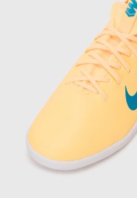Gul atletiksko med blåt Nike-logo, snøret op, og hvid struktureret sål på en ensfarvet grå baggrund.