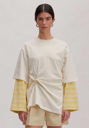 Frau mit dunklem Haar, die lagenweise creme- und gelb gestreifte Langarmshirts, ein cremefarbenes gedrehtes oversized Hemd und beige Shorts trägt.
