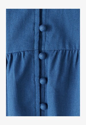 Sininen kangasvaate, jossa on nappi etuosa, jossa on teksturoidut pyöreät napit ja poimutettu vyötäröyksityiskohta. Materiaali näyttää kevyeltä ja teksturoidulta.