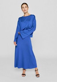 Blå langærmet kjole med en afslappet pasform, lavet af glat stof. Har brede ærmer og en svajende nederdel, parret med sorte stroppede sandaler.