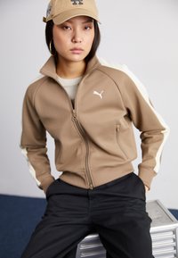 Brown zip-up jacket with cream accents, side pockets, and ribbed cuffs. Worn over a light-colored shirt, paired with black pants.