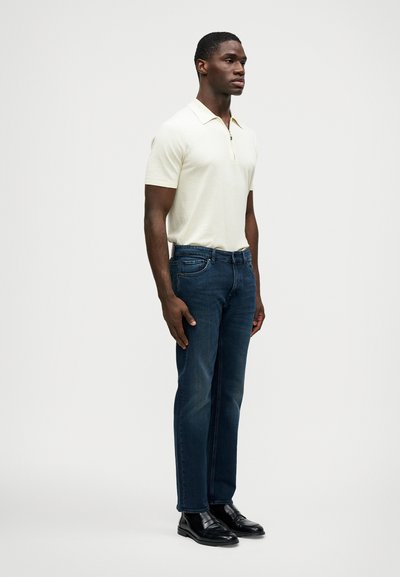 Homem veste uma polo de manga curta creme, jeans escuros e sapatos de couro pretos, de pé e virado ligeiramente para a direita, contra um fundo simples.