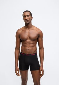 Melnas bokseršorti ar sarkanu "HUGO" jostas daļu, izgatavoti no gluda un elastīga materiāla, pieguloša dizaina un vidēja auguma garuma līdz augšstilbam.