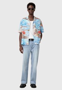 Kurzärmliges hellblaues Hawaii-Hemd mit roten und braunen Motiven, kombiniert mit hellen Jeans und schwarzen Schuhen. Mit Sonnenbrille accessoriert.