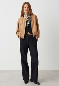Giacca bomber in suede color sabbia con zip frontale, top nero corto, pantaloni larghi neri e una sciarpa nera e bianca con motivo.