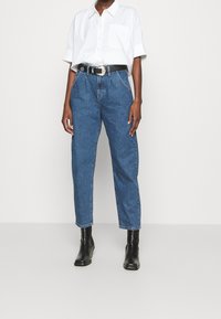 Camicetta bianca a maniche corte con taschino, abbinata a jeans blu a vita alta e una cintura nera. Gli stivaletti neri completano l'outfit.