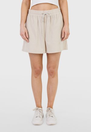 Short beige à cordon porté par une personne avec des baskets blanches et un haut court blanc, debout contre un fond uni.