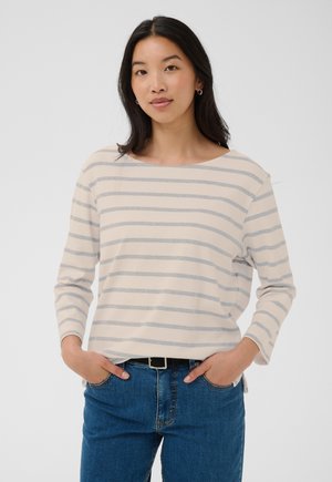 Jeune femme aux longs cheveux foncés, debout, les mains dans les poches, portant un haut crème à manches longues avec des rayures horizontales gris clair et un jean bleu.