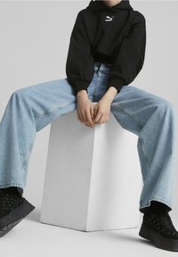 Felpa nera con logo bianco, jeans larghi blu chiaro e sneakers platform nere. Modello seduto con le gambe divaricate su un pedestal bianco.