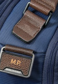 Donkerblauwe stoffen achtergrond met een bruine leren riem en label met geperste initialen "M.P." en merknaam "Piquadro" in zilverkleurig beslag.