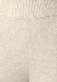 Tissu beige en maille avec une ceinture côtelée. La texture est douce et légèrement duveteuse, présentant un motif subtil et uniforme sur toute la surface.
