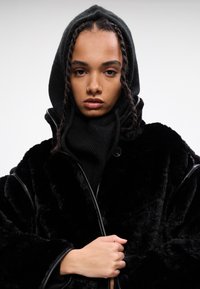 Abrigo negro de felpa con capucha, que presenta una superficie texturizada y ribetes en contraste. La modelo lleva una prenda ajustada con capucha de color oscuro debajo.