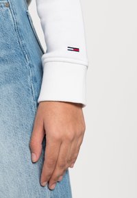 Chemise blanche à manches longues avec des poignets côtelés, ornée d'un petit logo bleu marine, rouge et blanc sur la manche, associée à un denim bleu clair.