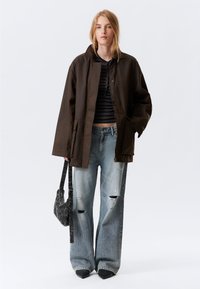 Brun oversized jakke, sort ribbet top, slidte lyseblå vidde bukser med vide ben, spidse sorte sko og en lille grå taske.