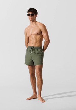 Uomo muscoloso che indossa pantaloncini da bagno verde scuro con cordino e occhiali da sole rettangolari neri, in piedi a piedi nudi su sfondo chiaro.