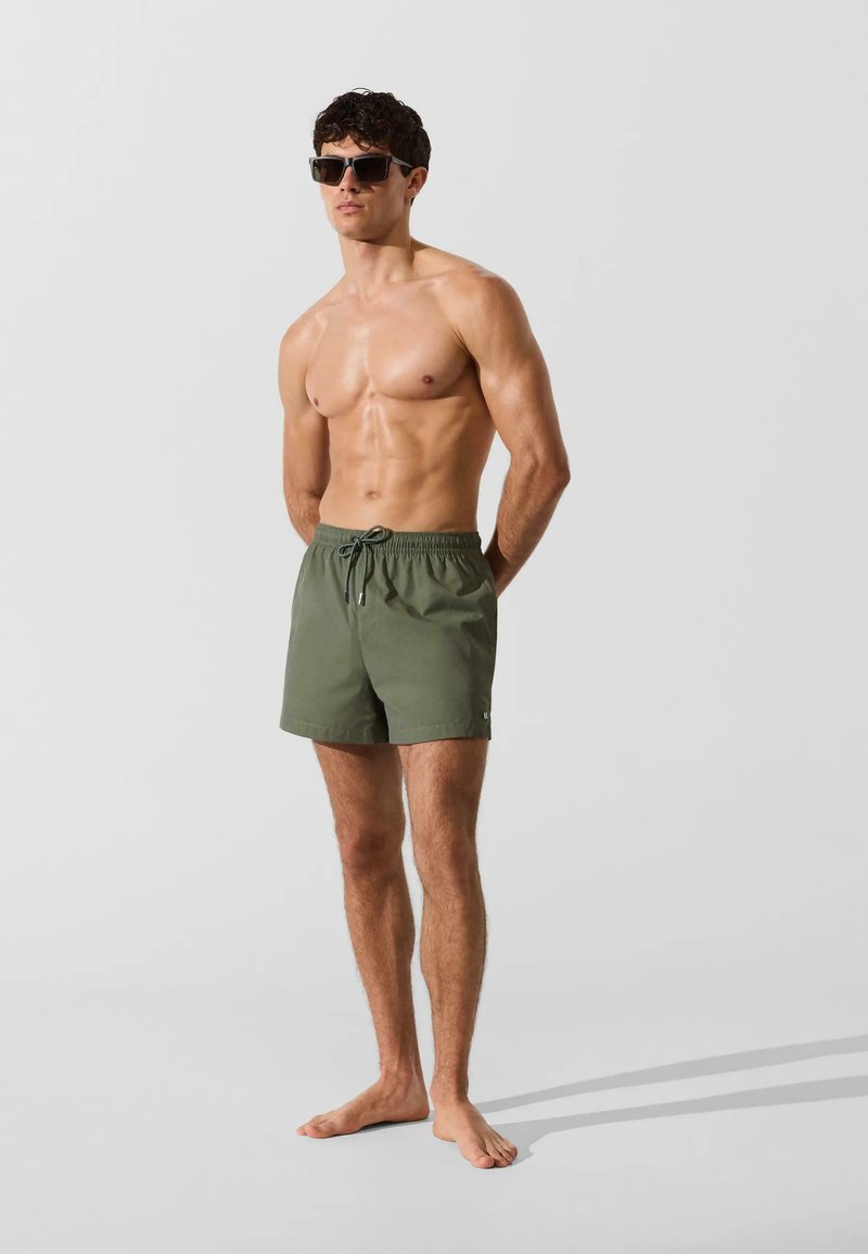 Homme musclé portant un short de bain vert foncé avec cordon et des lunettes de soleil rectangulaires noires, debout pieds nus sur un fond clair.