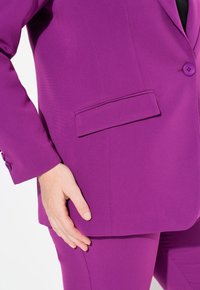 Fialový sako s jedním knoflíkem, detaily kapes a hladkou strukturou, v kombinaci s odpovídajícími kalhotami, které zdůrazňují konzistentní barvu a design.