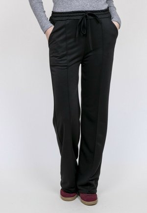 Pantalon noir à cordon de serrage avec coutures avant, porté avec un pull gris et des chaussures sans lacets bordeaux, mains dans les poches, debout sur fond blanc.
