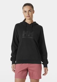 Sudadera negra con un bolsillo canguro delantero y capucha con cordón, que presenta un logotipo negro mate "HH" y el texto "HELLO HANSEN". Combinada con pantalones cortos rosas.
