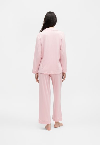 Γυναίκα στέκεται ξυπόλητη με την πλάτη γυρισμένη, φοράει ανοιχτό ροζ πυτζάμες με μακριά μανίκια και χαλαρό παντελόνι σε λευκό φόντο.