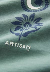 Tela verde con un diseño floral en azul oscuro y acentos, junto con la palabra "ARTISAN" en una fuente blanca y estilizada.
