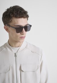 Gafas de sol con forma rectangular y lentes oscuros, combinadas con una chaqueta de cremallera color beige claro que presenta dos bolsillos frontales y una textura suave.