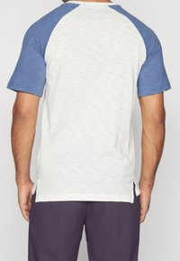 T-paita lyhythihaisella raglan-leikkauksella, jossa on vaaleanharmaa vartalo ja siniset hihat, pehmeä rakenne ja sivuhalkiot liikkumisen helpottamiseksi.