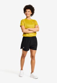 Gul kortärmad idrottströja med struktur, tillsammans med svarta shorts med resårmidja och logotyp. Vita sneakers fulländar outfiten.